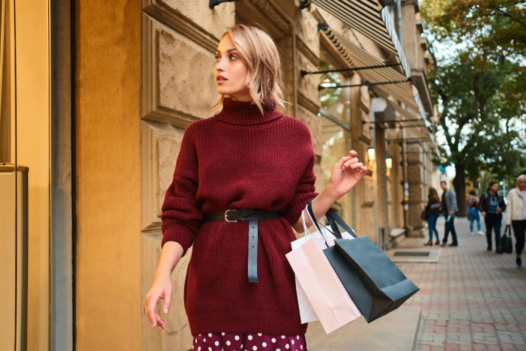 Une femme qui porte une robe pull avec une ceinture et qui tient des sacs 